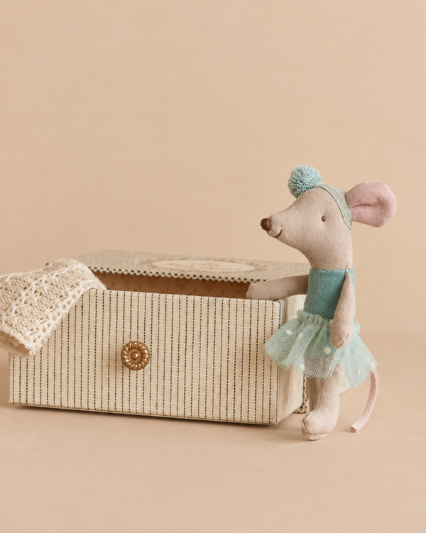 A Maileg Dance Mouse in Daybed, Little Sister - Mint stands beside a striped rectangular box with a knit blanket draped inside.