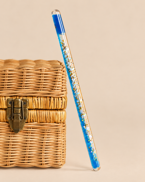 A clear liquid-filled Konges Sloejd Magical Glitter Wand - Rocket containing blue sequins and rocket-shaped glitter, leaning against a woven basket.
