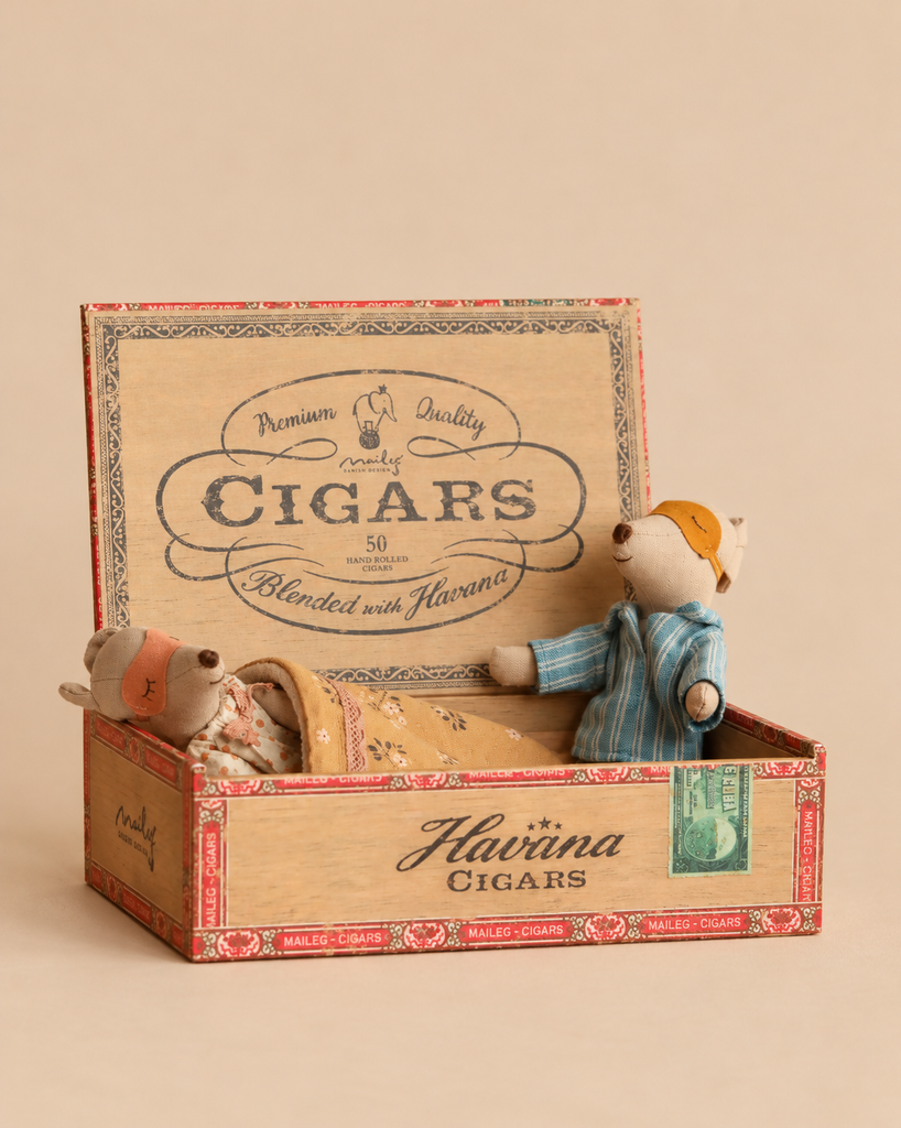 Two small stuffed bears in pajamas inside a Maileg Family Breakfast Set cigar box, with one bear lying under a blanket and the other standing.
