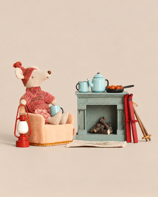 A Maileg Winter Chalet Set featuring a winter ski mouse in a knit sweater and hat sitting in an armchair with a mug, beside a fireplace with a tea set, food, a red lantern, and a pair of skis.