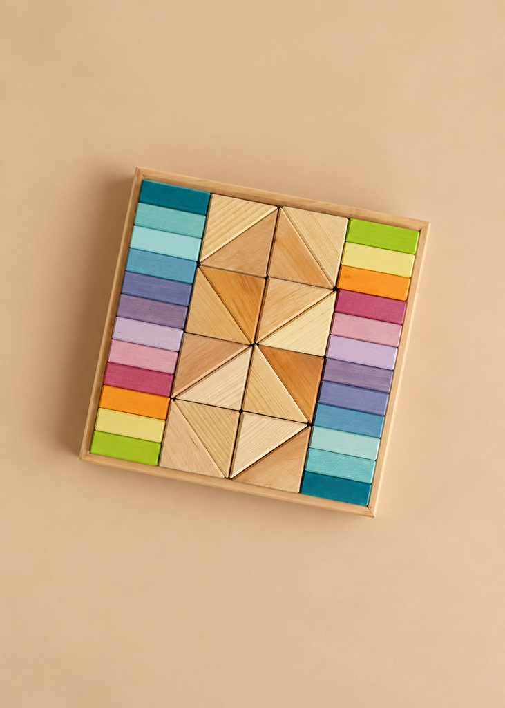 A wooden square tray from the Pastel Duo Block Set contains a central cluster of natural wood triangles flanked by columns of pastel building blocks in rainbow gradients.