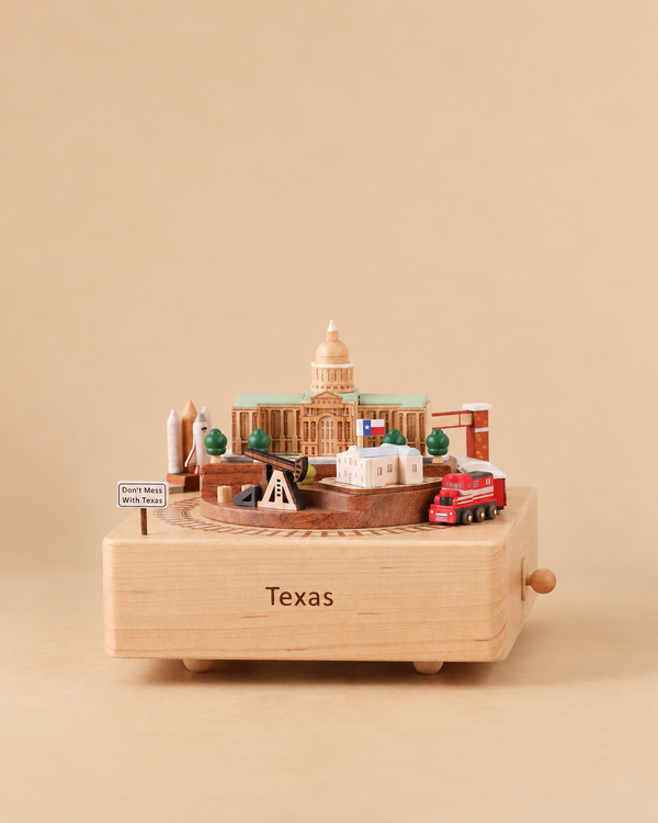 A Texas Wooden Music Box featuring a rotating platform with miniature models of the Texas State Capitol, the Alamo, an oil derrick, a rocket, and a red truck, complete with a sign reading “Don’t Mess With Texas.”