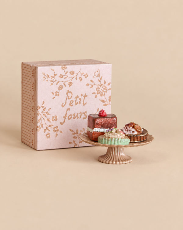Three Maileg Miniature Cakes & Cake Stand pastries displayed on a small pedestal stand in front of a pink box labeled “Petit fours.”