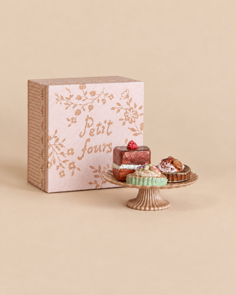 Three Maileg Miniature Cakes & Cake Stand pastries displayed on a small pedestal stand in front of a pink box labeled “Petit fours.”