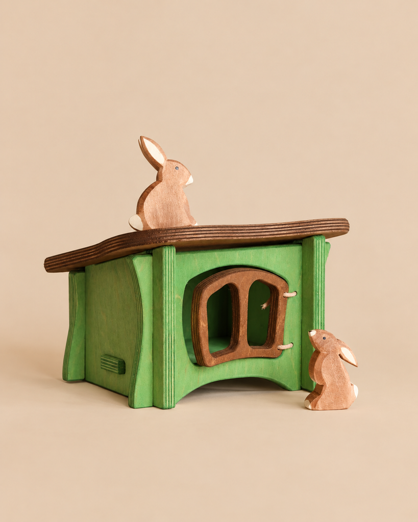 An Ostheimer Rabbit Hutch in green wood with a brown arched door, featuring one wooden rabbit sitting on the roof and another standing beside the handcrafted structure.