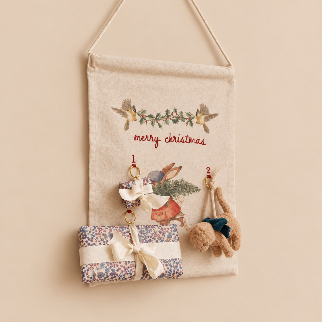 Hanging organic cotton Christmas Advent Calendar decorated with a rabbit illustration, holly, and “merry christmas,” featuring numbered rings holding small gift-wrapped packages and a plush animal toy.