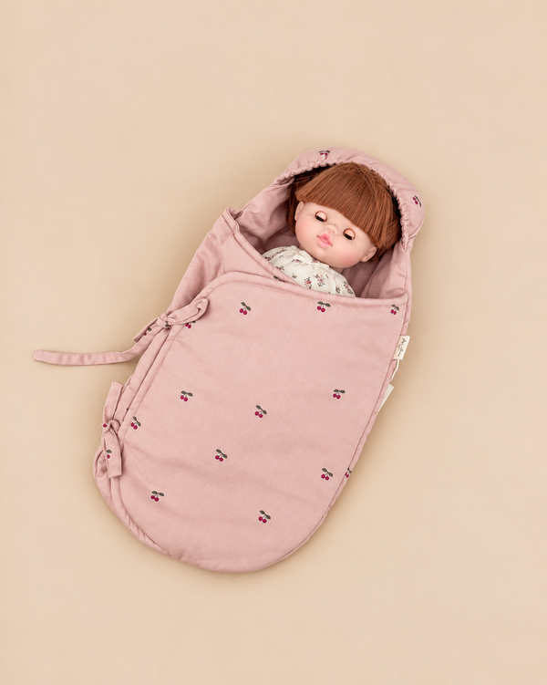 A doll tucked into a Konges Sloejd Doll Sleeping Bag - Cherry Blush made of organic cotton with a small cherry pattern.