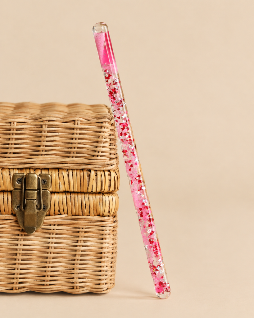 A Konges Sloejd Magical Glitter Wand - Pink Mix filled with liquid, pink glitter, and star-shaped confetti leaning against a small wicker box.