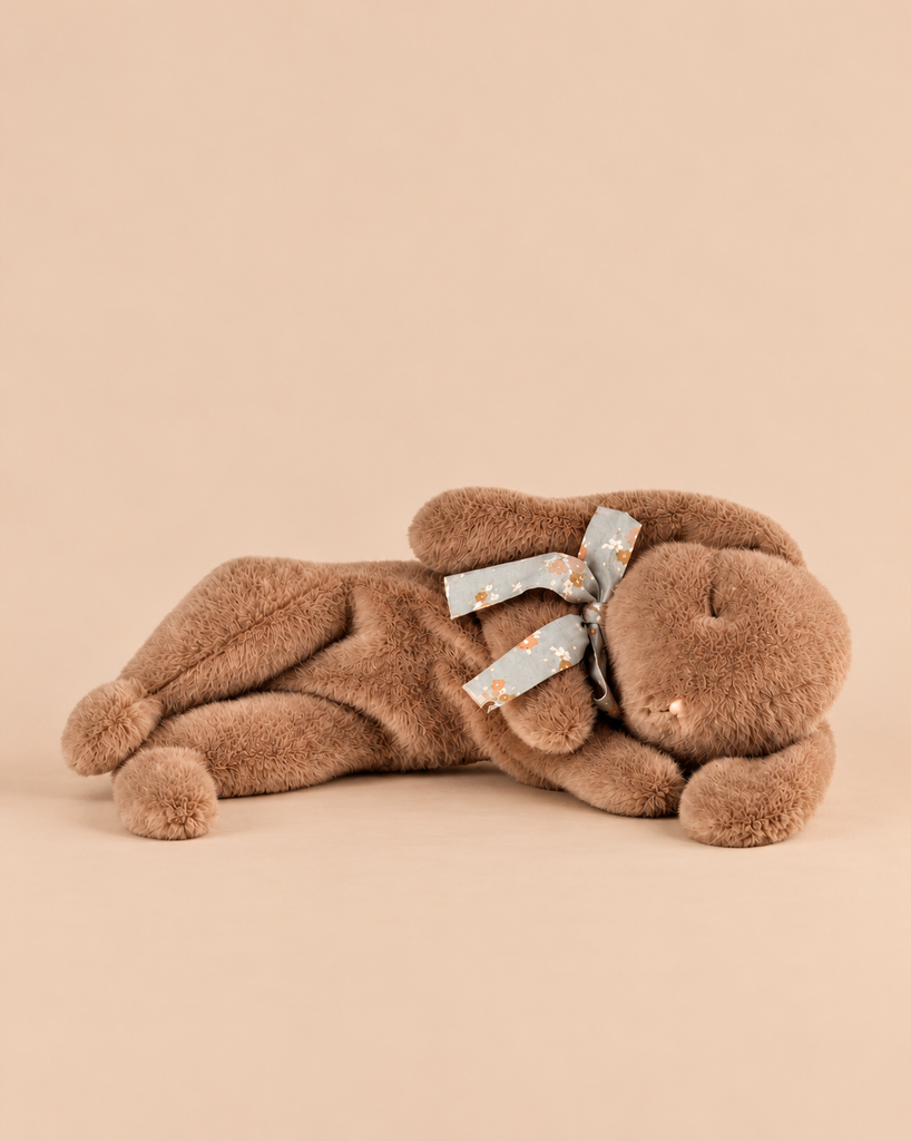 A soft brown Maileg Plush Sleeping Bunny - Soft Brown lies on its side, wearing a small floral ribbon around its neck.
