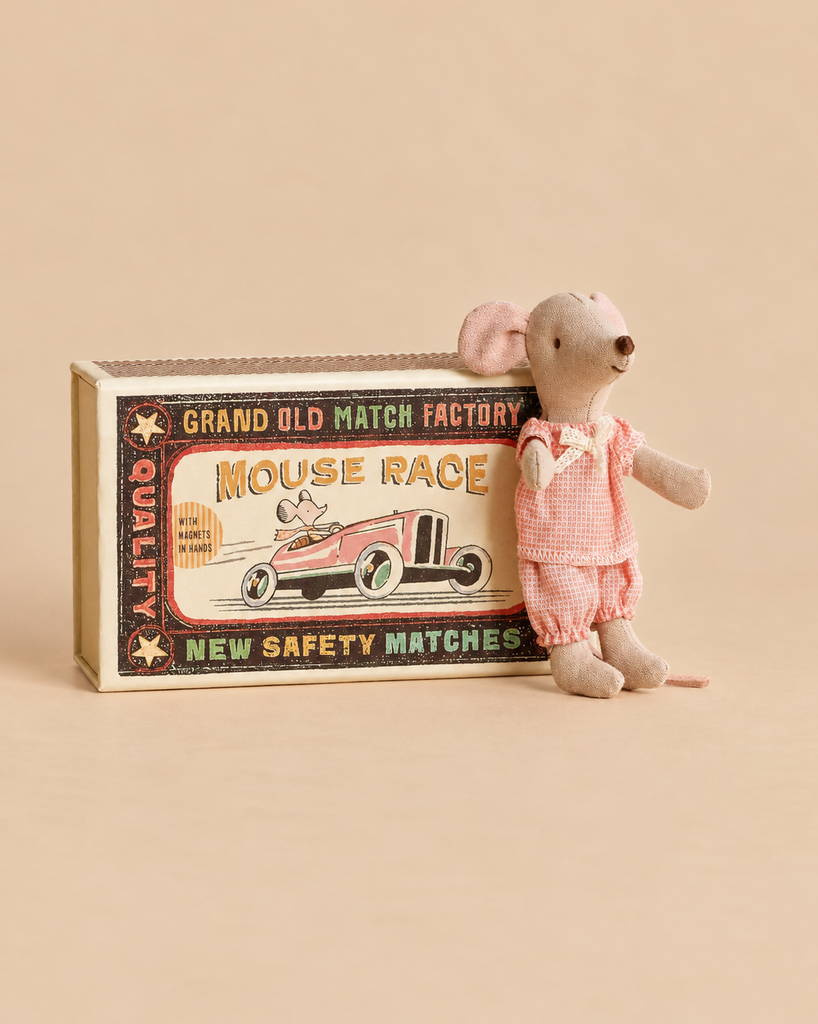 A small Maileg Little Sister’s Kitchen Set mouse in pink pajamas standing next to a matchbox-style toy box featuring an illustration of a mouse in a race car.