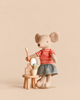 A Maileg Big Sister Getting Ready Set mouse in a striped shirt and skirt stands beside a table and stool, holding a toothbrush, a small bottle, and a tube of cream in front of a tiny mirror.