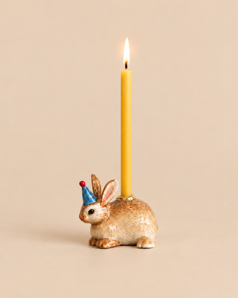 A Bunny Rabbit Cake Topper - Final Sale featuring a ceramic bunny with gold accents and a blue party hat, serving as a porcelain holder for a lit yellow taper candle.