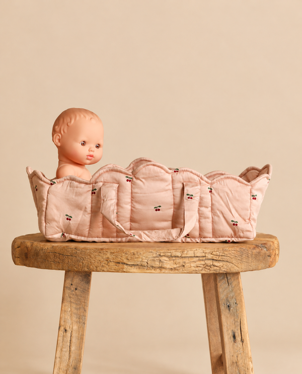 A toy doll sits inside a pale pink quilted Konges Sloejd Doll Lift - Cherry Blush with a cherry print, resting on a rustic wooden stool.