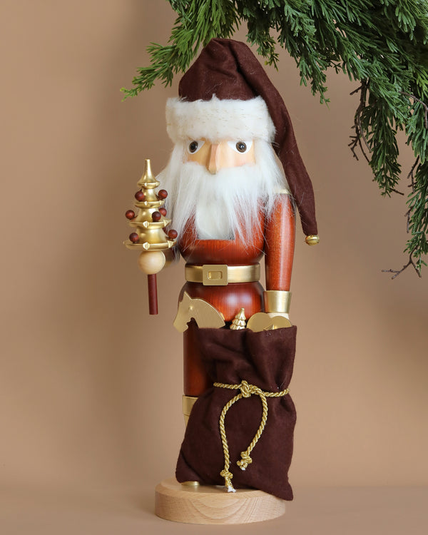 This limited-edition Christian Ulbricht Collectible Wooden Nutcracker - Natural and Gold Santa showcases a white-bearded Santa wearing a brown hat and holding a decorative golden tree. It is accompanied by a brown sack tied with a gold rope, while greenery elegantly drapes from the top right corner.