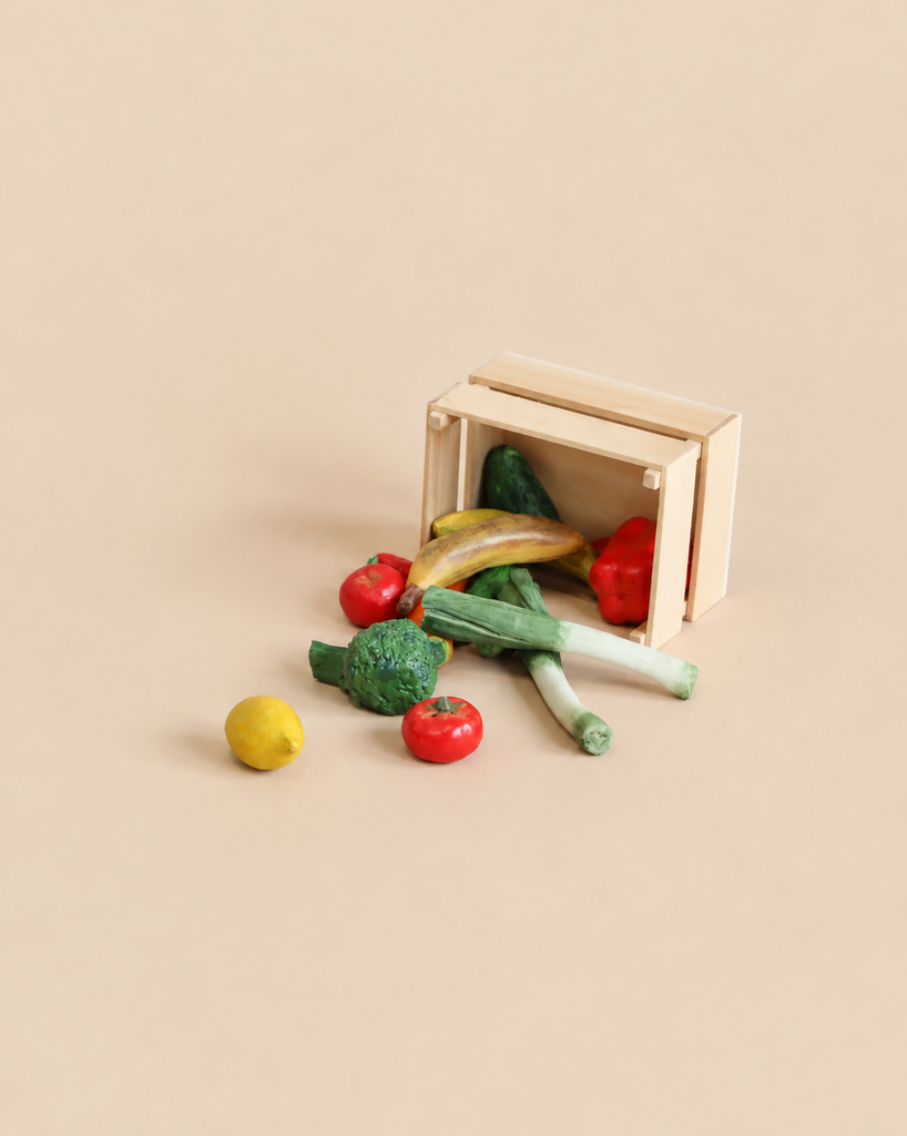 A wooden crate from the Maileg Grocery Wagon Set tipped on its side, spilling miniature fruits and veggies like broccoli, leeks, a banana, and tomatoes.