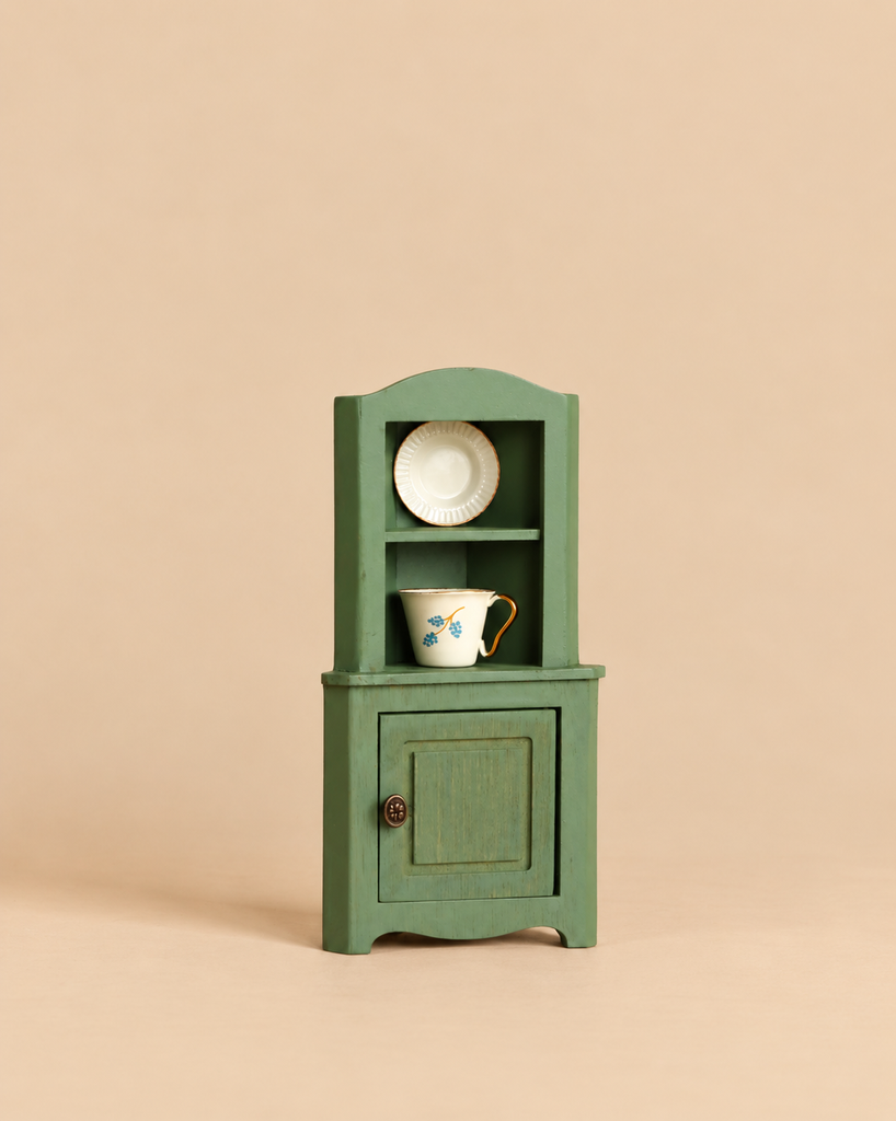 Maileg Corner Cabinet, Mouse Size - Dark Green miniature corner cabinet with a vintage finish, featuring two open shelves containing a small plate and cup above a single cupboard door.