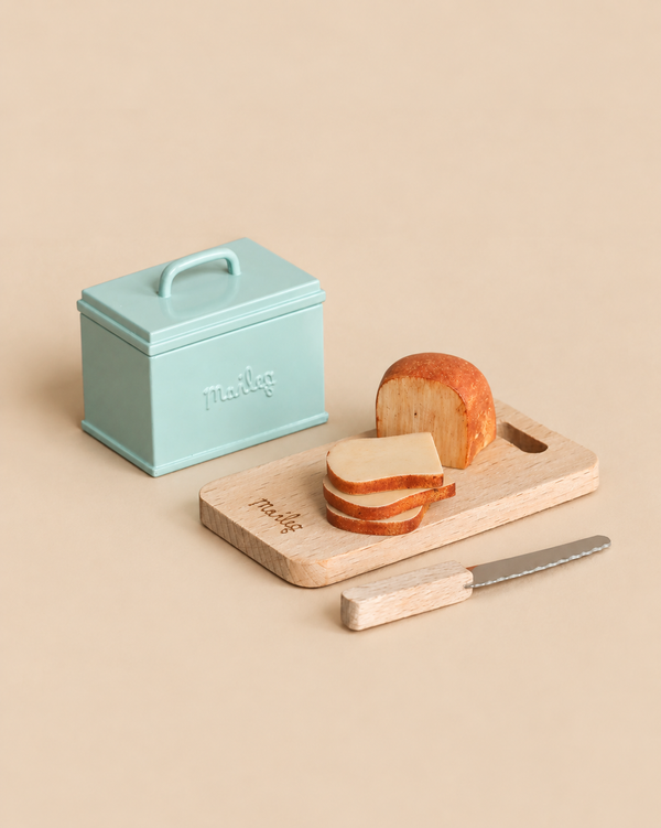 A small turquoise metal Maileg Miniature Bread Box sits next to a wooden cutting board with a toy loaf of bread, three slices, and a serrated knife from a miniature kitchen playset.