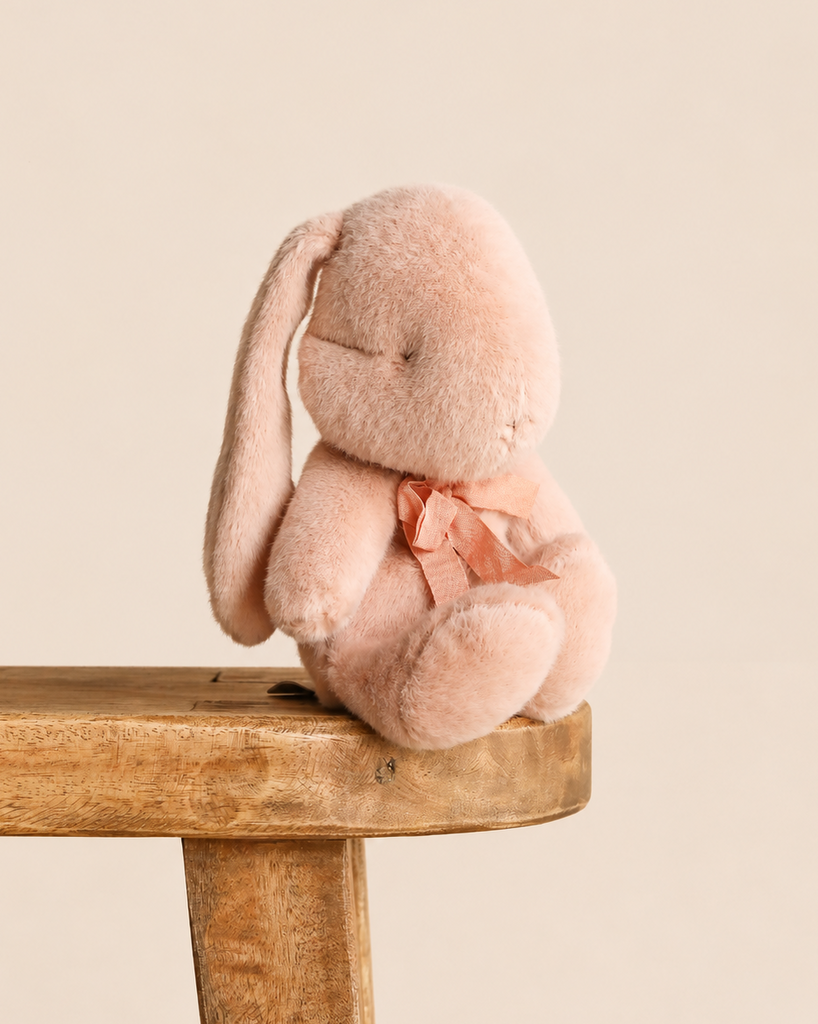 A small, pale pink Maileg Plush Bunny, Small - Powder with one long drooping ear and a tied ribbon at its neck, sitting on a wooden stool as a soft toy.