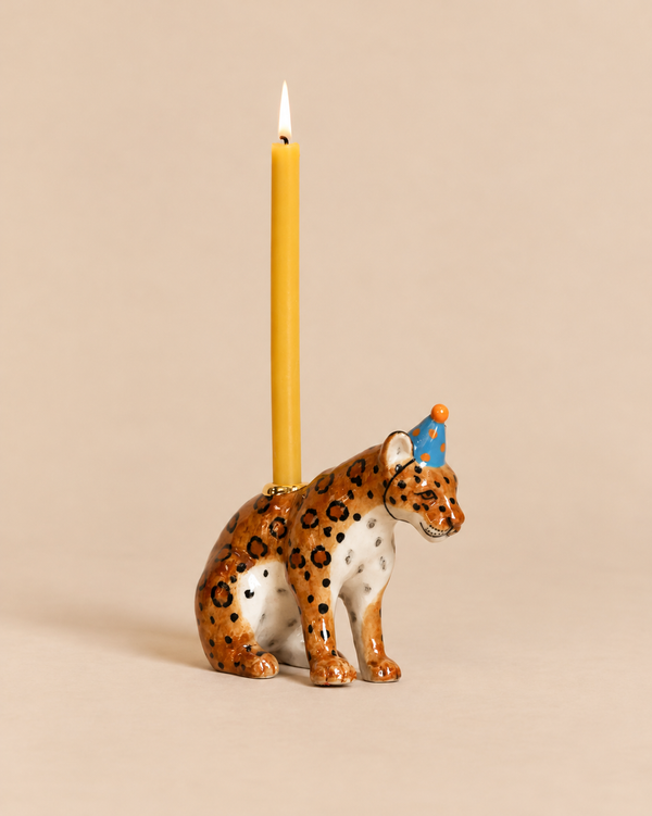 A Jaguar Cake Topper - Final Sale featuring a ceramic jaguar figurine in a blue polka-dot party hat, with a candle holder on its back holding a lit yellow taper candle.