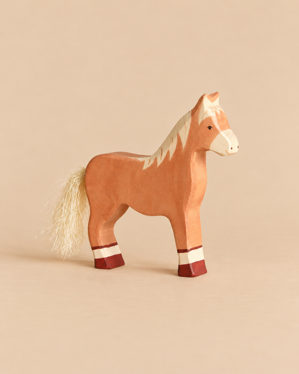 A handcrafted Holztiger Horse, Standing, Light Brown wooden toy with a light brown finish, a white mane, a string tail, and painted dark red markings on its lower legs.