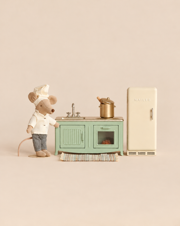 A Maileg Kitchen Set with Chef Mouse featuring a stuffed toy chef standing next to a miniature green kitchen unit, a sink, a gold pot, and a cream-colored refrigerator.