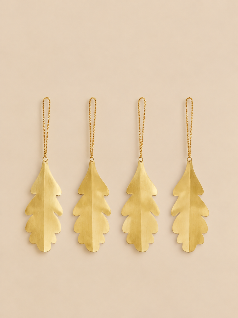 Four gold-colored brass Ferm Living Oak Leaf Ornaments - Set of 4 - Brass, each shaped like an oak leaf and hanging from a thin chain.