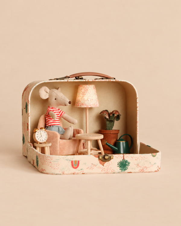 A small Maileg mouse in a striped shirt sits in an armchair inside a Maileg On The Go Suitcase Set, which also contains miniature dollhouse furniture, a lamp, table, potted plant, watering can, and alarm clock.