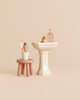 Maileg Powder Room Set featuring a miniature bathroom sink with a gold faucet and perfume bottle, next to a stool set holding a cup, toothbrush, and toiletry bottle.