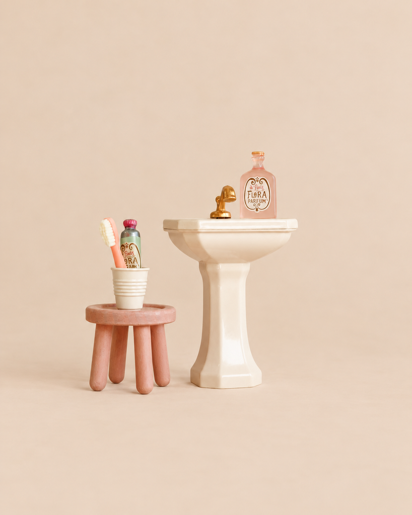 Maileg Powder Room Set featuring a miniature bathroom sink with a gold faucet and perfume bottle, next to a stool set holding a cup, toothbrush, and toiletry bottle.