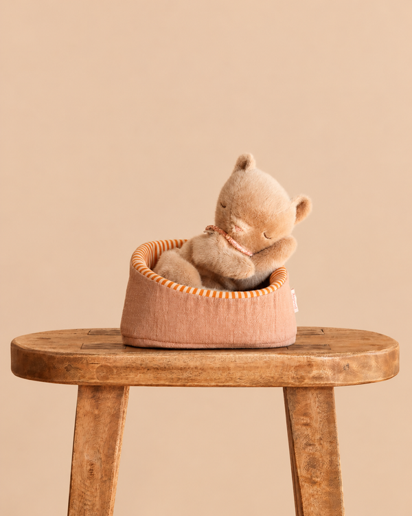 A Maileg Small Sleeping Kitten in Basket resting in a small fabric basket placed on a wooden stool.