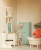 A stuffed toy mouse stands at a sink in a tiny bathroom, with dollhouse furniture like a toilet, bathtub, and green cabinet—perfect for imaginative play with the Maileg Fully Furnished Dollhouse Set + Surprise Gift.