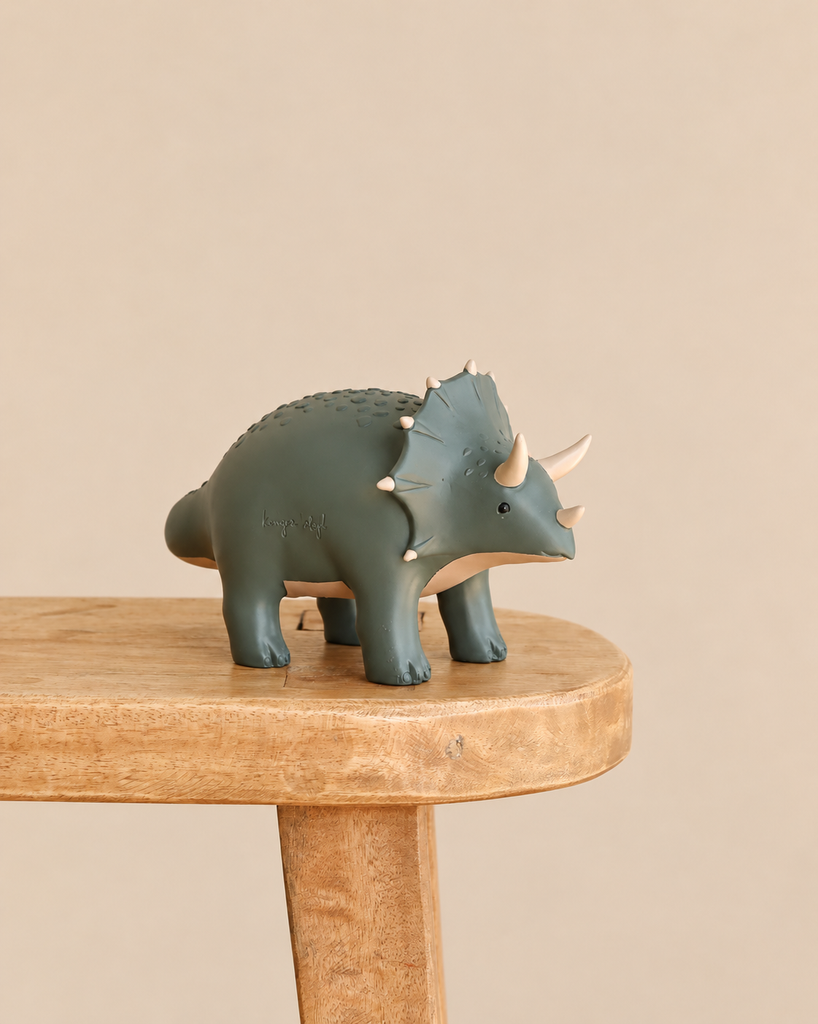 Konges Sloejd Money Bank - Dino, a dark green triceratops with cream-colored horns standing on a wooden stool.