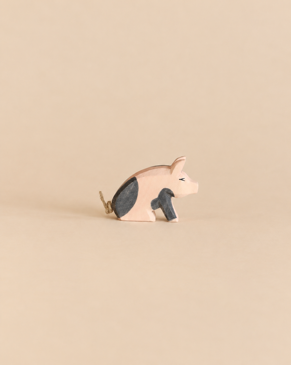 Ostheimer Spotted Piglet - Sitting, a handcrafted wooden toy in a sitting position with black patches and a simple twine tail, perfect for imaginative play.