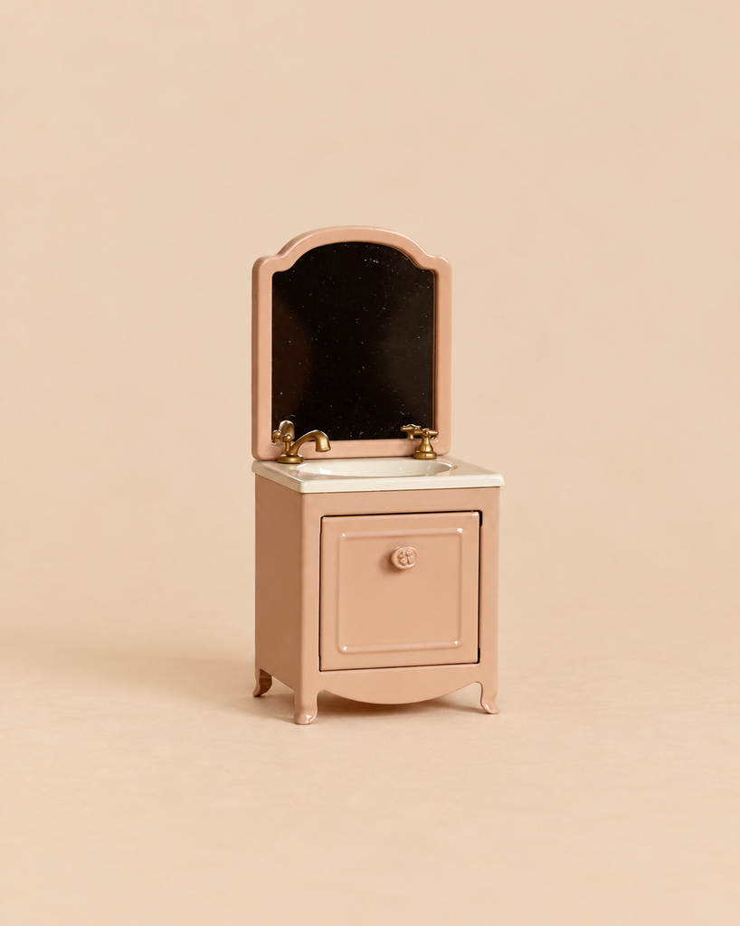 Maileg Sink Dresser With Mirror, Mouse - Dark Powder, a miniature pink bathroom vanity featuring a sink, two gold-toned faucets, and an attached mirror.