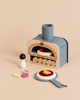 A Wooden Make Me A Pizza Set featuring a wooden toy pizza oven, pepper grinder, and pizza toppings on a plain surface for pretend play.