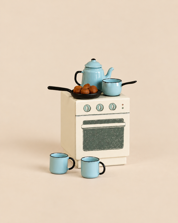 A Maileg Madam Blue’s Favorites toy kitchen stove topped with a blue teapot, a small saucepan, and a frying pan containing small brown treats, with two blue mugs on the floor in front.