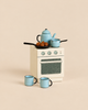 A Maileg Madam Blue’s Favorites toy kitchen stove topped with a blue teapot, a small saucepan, and a frying pan containing small brown treats, with two blue mugs on the floor in front.