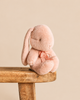 A small, light pink Maileg Plush Bunny, Small - Powder with one long drooping ear and a matching ribbon bow around its neck sits on a wooden stool.