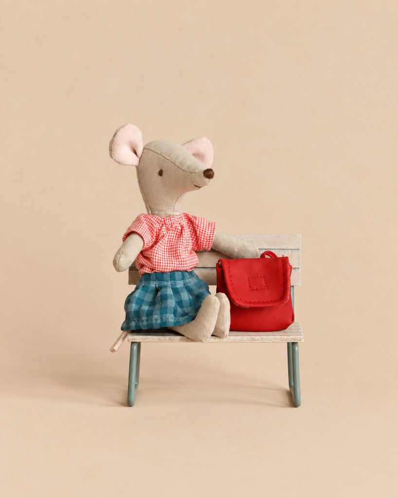 Maileg Big Sister With Backpack - Red mouse in a red gingham shirt and blue plaid skirt sitting on a small bench next to a red backpack.