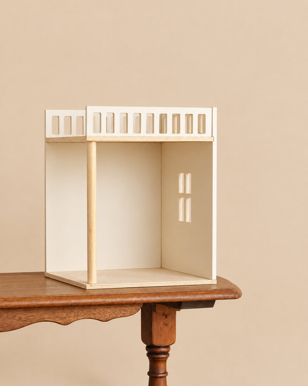 A Maileg House of Miniature Bonus Room - Bedroom, featuring a small, open-fronted wooden room with a balcony railing, resting on a wooden table as a dollhouse accessory.