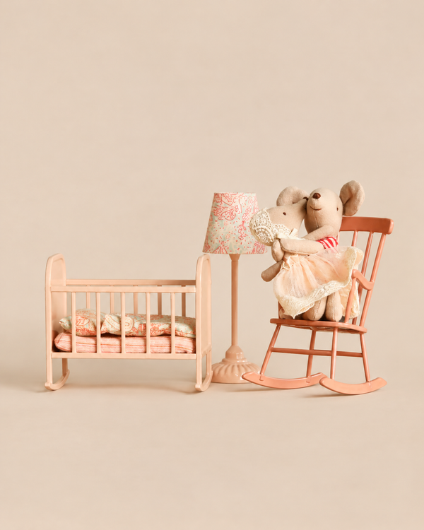 Two Maileg Big Sister and Me Nursery Set mice sit in a pink rocking chair next to a miniature cradle and a small table lamp.