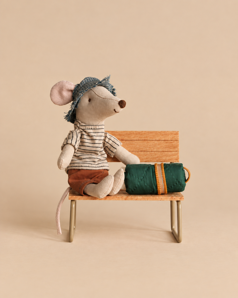 A Maileg Hiker Mouse With Brown Striped Shirt - Big Brother wearing brown shorts and a small hat sits on a miniature wooden bench next to a green sleeping bag for its hiking accessories.