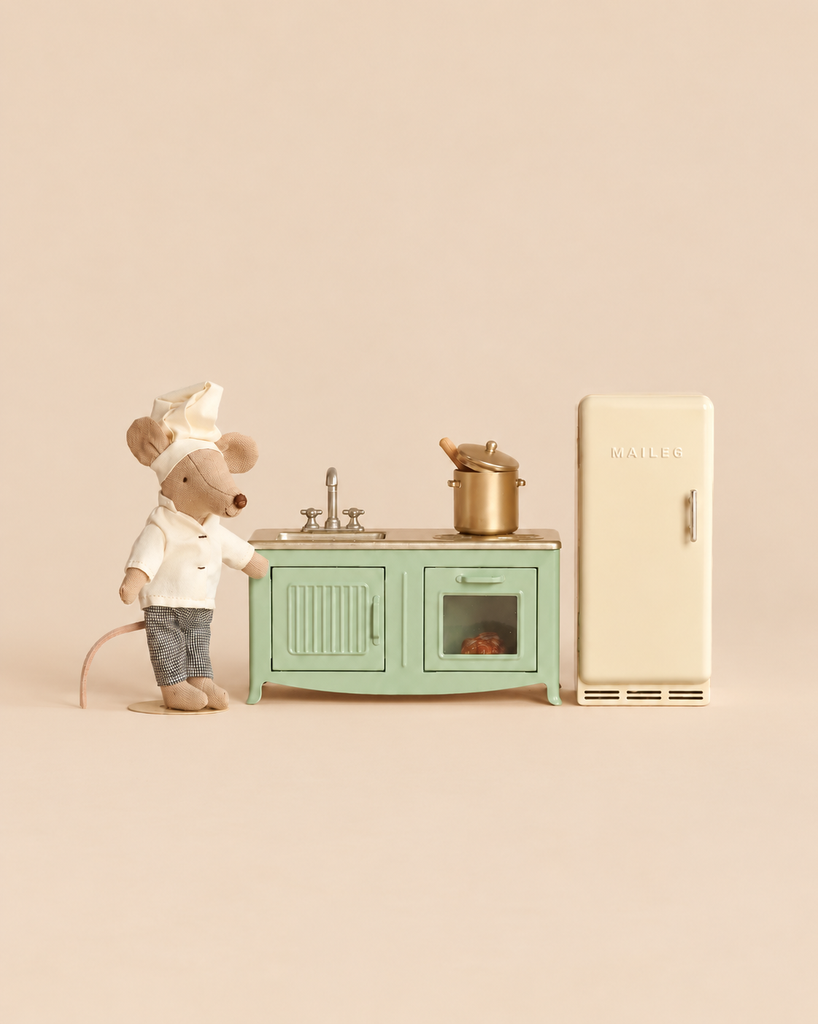 A Maileg Kitchen Set with Chef Mouse features a chef mouse toy standing next to a miniature kitchen with a green stove, a pot, a sink, and a cream-colored refrigerator.