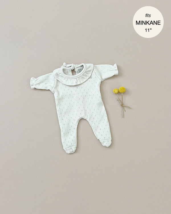 A light-colored, long-sleeve footed onesie with a ruffled collar, tailored for 11" dolls, is laid out on a beige background. Two yellow billy balls are placed beside it. A circular label reads "fits MINIKANE 11''", highlighting its suitability for Minikane Doll Clothing | Sleep Well Dotted Linen made in France.