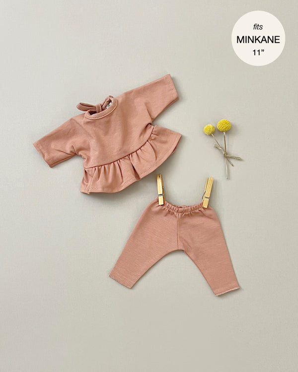 A Minikane Clothing | Ophélia Set in Brown Sugar for an 11" Minikane Babies doll is displayed. The cotton hand-washable outfit includes a peach-colored long-sleeve top with a ruffled hem and matching pants, both secured by wooden pegs. Two yellow billy ball flowers are placed beside the outfit against a light background.