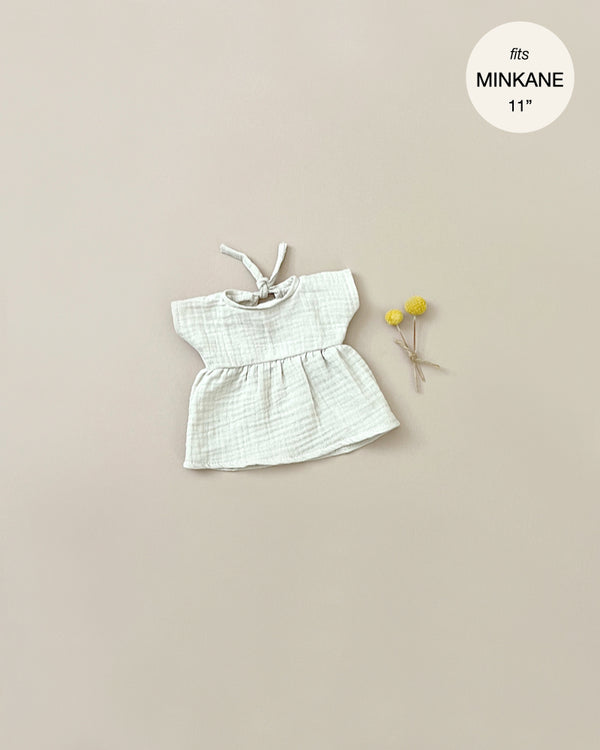 A Minikane Doll Clothing | Faustine Dress and Bonnet Set is laid flat on a beige background. Beside it, a small bundle of two yellow flowers adds a touch of charm. A label in the top right corner reads "fits Minikane Babies 11".