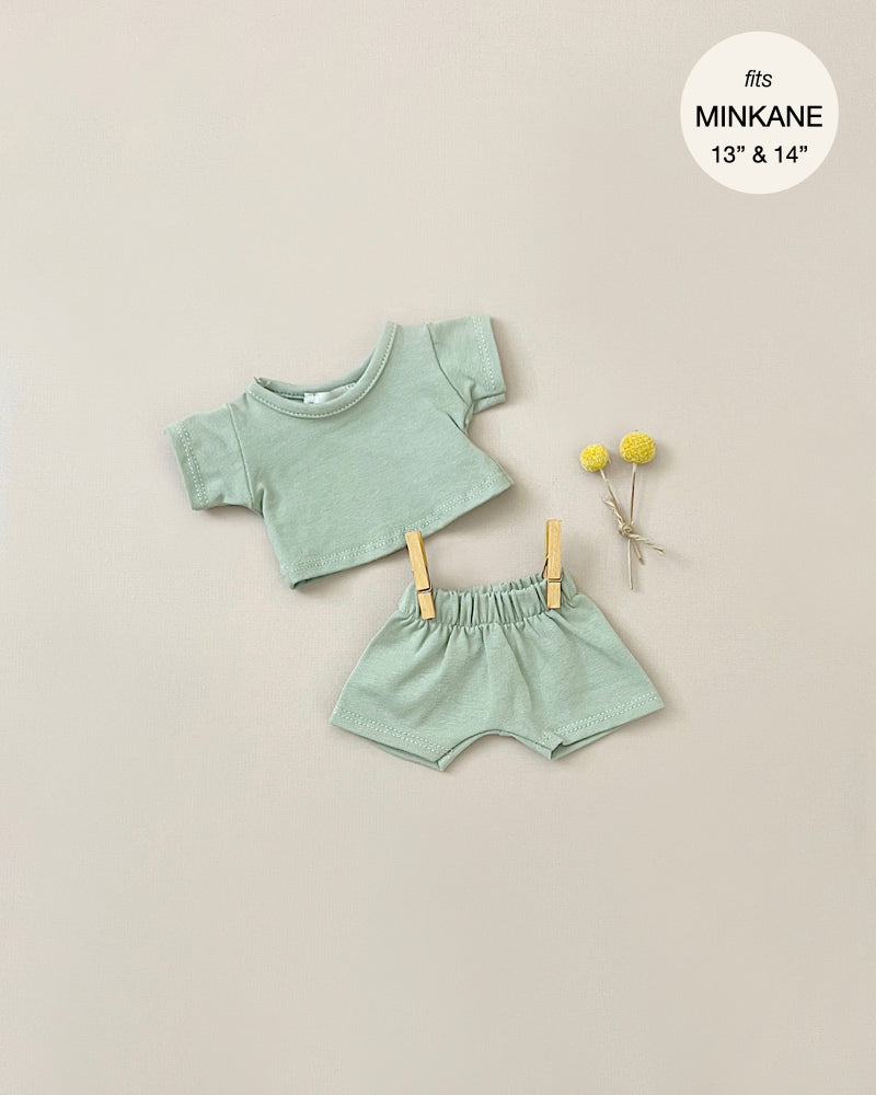 A tiny Minikane Doll Clothing | Vito T-Shirt and Shorts Set in Green consisting of a short-sleeved crop top and matching bottoms is neatly displayed. Two small yellow flowers are placed beside the outfit. There's text that reads "fits Minikane Gordis dolls 13" & 14".