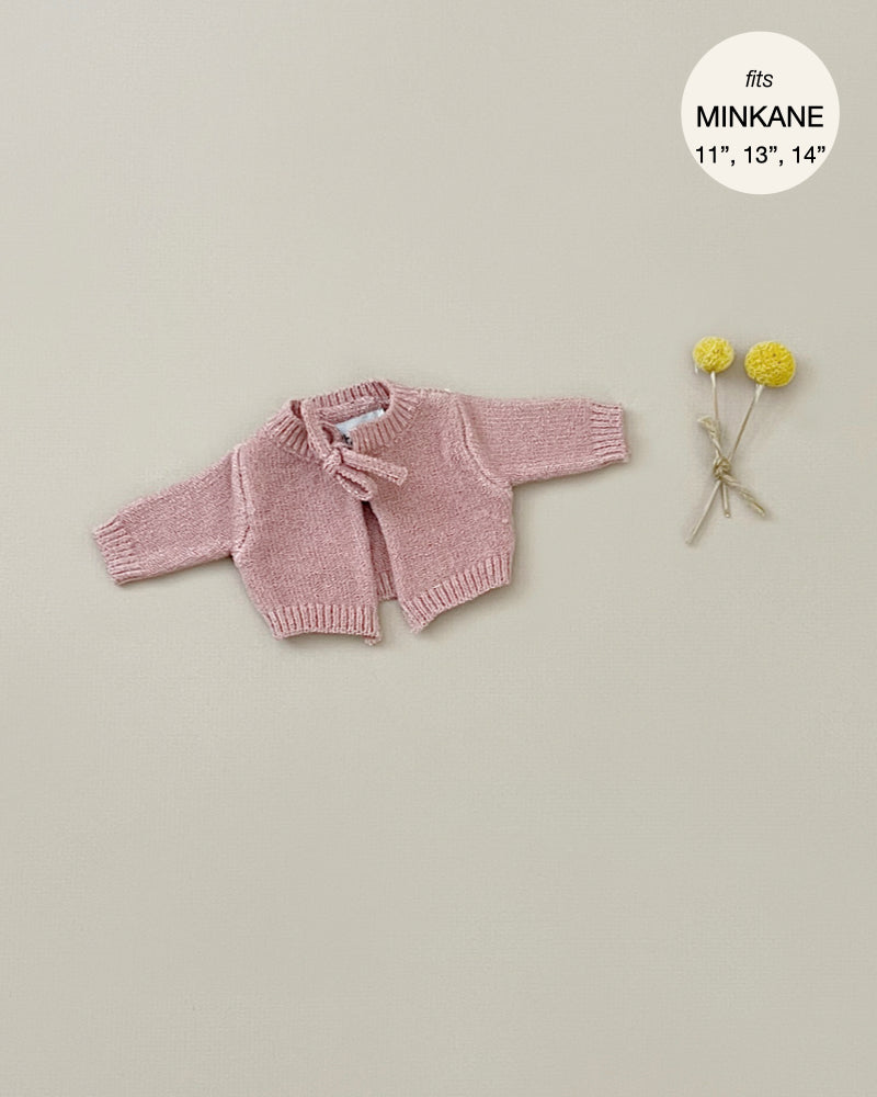 A small, pink knitted Minikane Doll Clothing | Tea Pink Knit Alix Cardigan with long sleeves and a ribbon closure is laid out on a beige background. To the right, two yellow billy buttons (craspedia flowers) are tied together with a thin, neutral-colored ribbon. The text in the corner reads "fits Minikane Babies 11'', 13'', 14''." Hand washable.