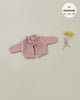 A small, pink knitted Minikane Doll Clothing | Tea Pink Knit Alix Cardigan with long sleeves and a ribbon closure is laid out on a beige background. To the right, two yellow billy buttons (craspedia flowers) are tied together with a thin, neutral-colored ribbon. The text in the corner reads "fits Minikane Babies 11'', 13'', 14''." Hand washable.