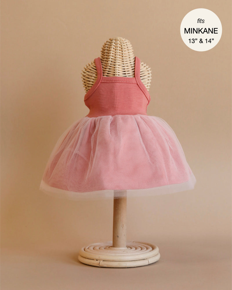 A child's dress displayed on a wicker dress form, perfect for Minikane dolls. The Rosella Tutu in Marsala features a sleeveless pink bodice and a voluminous soft pink tulle skirt. Text in the top right corner reads, "fits 13” & 14”." The background is a plain, neutral color, showcasing its designed-in-France elegance.